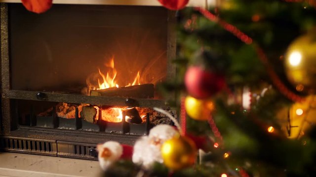 4k video of fire burning in fireplace next to decorated Christmas tree with glowing colorful lights. Perfect shot for winter celebrations and holidays