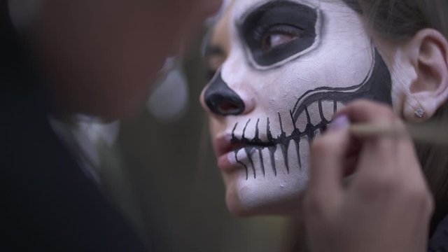 Woman's hands applying black paint on the face of the girl using the small brush close up. Young people preparing for the celebration of Halloween, painting the skull on the face. Professional makeup