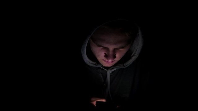 4K Man on a smart phone with light shining on his face. A smiling hooded teenager on a mobile device using a thumb to connect. A dark black background in shadow using a mobile swipe device.