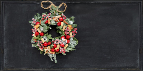 Christmas wreath made of fir branches, dried apples, cinnamon, red berries, bottle caps, red balls hanging on a black chalk board.