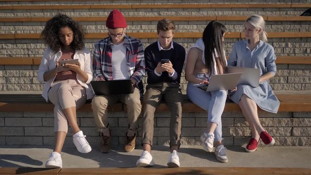 Multi-ethnic students sitting in a row absorbed by phones, computers, tabs. Device addiction.
