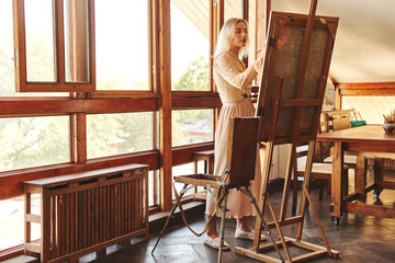 Fototapeta na wymiar Beautiful talented young woman artist paints a picture with brush and oil on an easel with palette in hand in stylish creative art studio