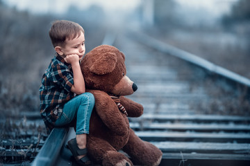 A little boy with a big teddy bear sits on the rails of the railway. Loneliness and sadness of a...