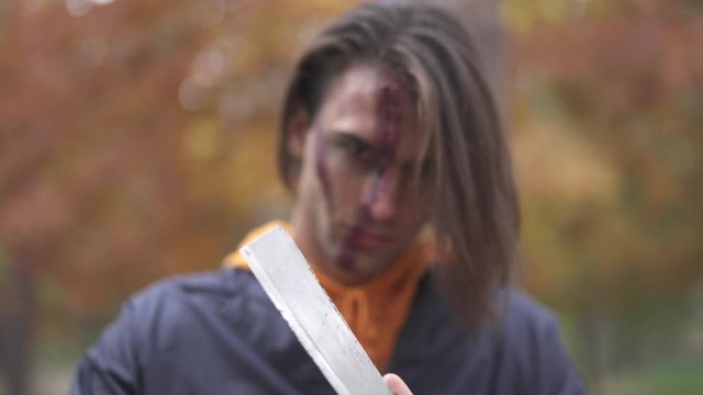 Handsome man with big ugly scar on his face demonstrating two cleavers, rubbing them on each other. The guy with Halloween makeup on face rubbing his knives with an evil look. Focus changing