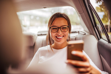 Portrait of a happy young woman using smart phone on a backseat of a taxi.