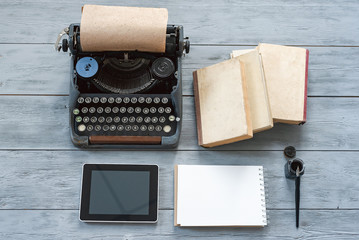 Typewriter, blank page notepad, stack of books and modern digital tablet on gray wooden background.