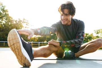 Photo of young caucasian man sitting on mat and stretching his leg