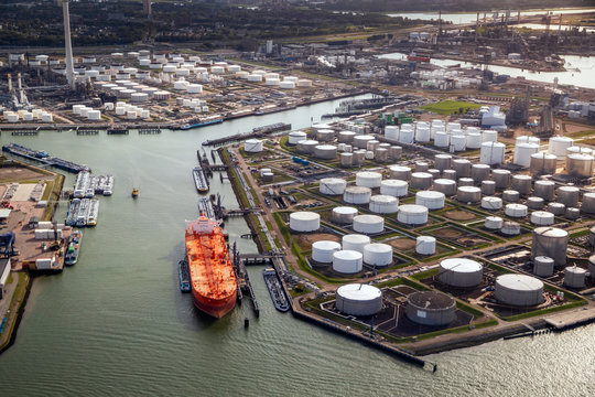 Aerial view of a large orange oil tanker moored at an oil storage silo terminal in an industrial port.
