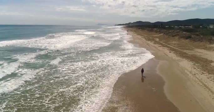 Vista de p&aacute;jaro de dron de playa dorada y extensa con una chica paseando con sombrero