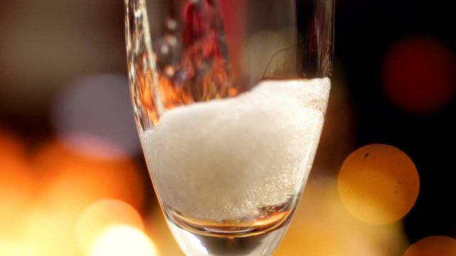 Macro slow motion video of pouring champagne from bottle in glass against glowing colorful lights. Gas bubbles and foam rising up. Perfect shot for parties,celebrations and holidays
