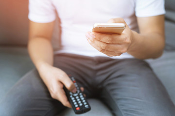 young man using television remote control and mobile smart phone  is sitting on a sofa. on or off...