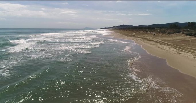 Dron vuela encima de las olas en playa de arena dorada y una pareja pasea