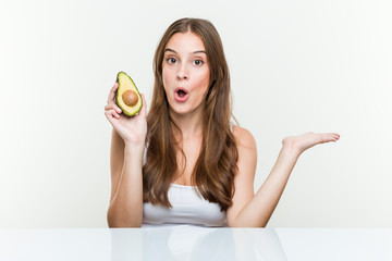 Young caucasian woman holding an avocado impressed holding copy space on palm.