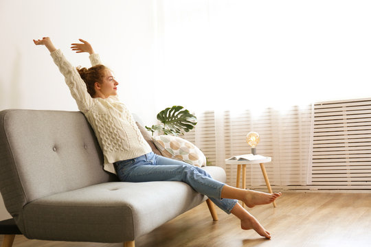 Young beautiful woman wearing white sweater on grey textile sofa at home. Attractive slim female in domestic situation, resting on couch in her lofty apartment. Background, copy space, close up.