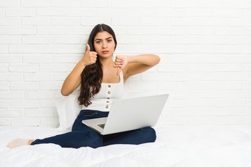 Young arab woman working with her laptop on the bed showing thumbs up and thumbs down, difficult choose concept