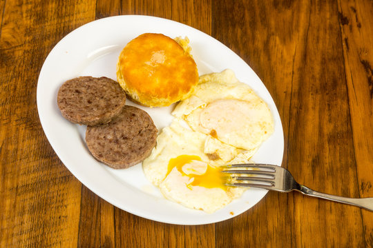 Overhead View Of Eggs Over Easy With Sausage And Buttered Biscuit On Rustic Wood Table