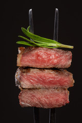 Three steak cuts or pieces on a metal fork with back light with rosemary closeup studio shot on black background isolated.