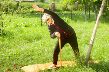 An elderly woman of 90 years old, with gray hair and a headband, doing fitness in the garden.