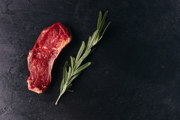 Raw beef meat piece steak with rosemary on black background. Top view, flat lay, place for text.