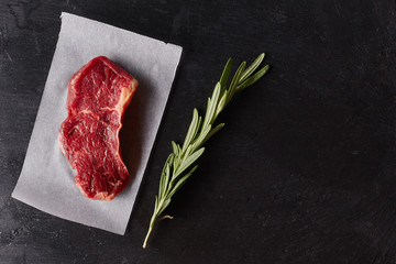 Raw beef meat piece steak on a paper with rosemary on black background. Top view, flat lay, place for text.