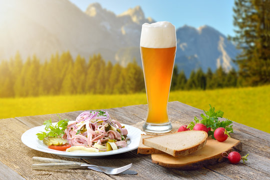 Deftiger Schweizer Wurstsalat mit einem Weizenbier draußen serviert, die Zugspitze im Hintergrund – Outside served sausage salad with a wheat beer, in the background the European alps