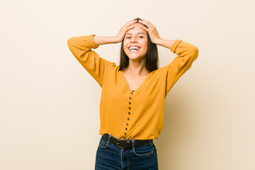 Young hispanic woman against a beige background laughs joyfully keeping hands on head. Happiness concept.