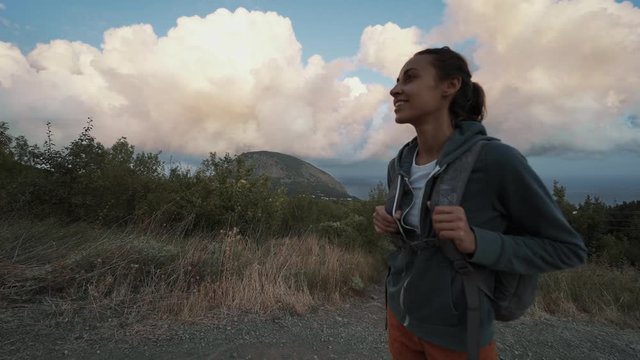 woman hiking mountains looking beautiful colorful amazing sunset sky. female hiker with backpack standind on mountain trail at seaside overlooking of amazing big rose clouds on blue sky over seaside