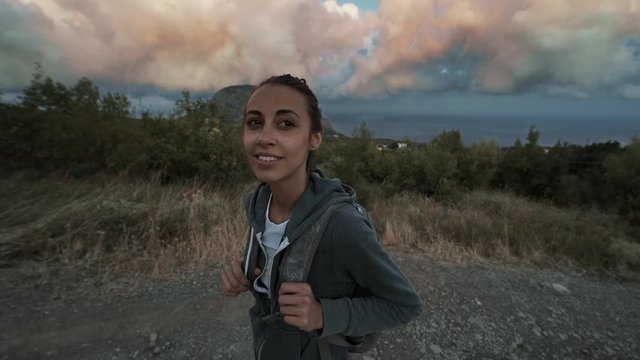 portrait of woman hiker looking beautiful colorful amazing sunset sky. female hiker with backpack standind on mountain slope at seaside overlooking of amazing big rose clouds on blue sky over seaside