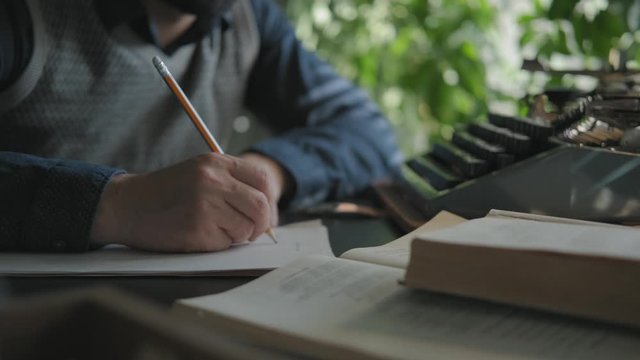 Male writer works sitting at the table. Medium shot: hands, a sheet of paper and a pencil. The writer is working on a draft, writes in pencil. A man writes on paper, typewriter is in front of him