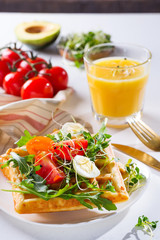 Fresh baked sweet belgian waffles with eggs, tomatoes, micro green and avocado served on a plate with orange juice on marble table. Healthy breakfast