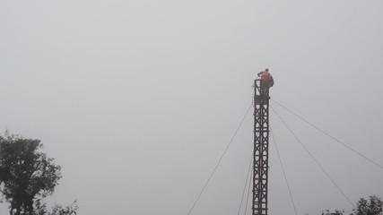 Workers in an orange vest and wearing safety harnesses working on a tower, telephone pole supported by wires and surrounded by fog