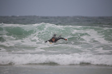 Young active man in black swimsuit lying on surfboard and swimming in water with waves on gloomy day