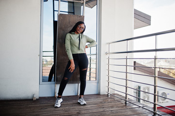 Fototapeta na wymiar City portrait of positive young dark skinned female wearing green hoody and eyeglasses standing at balkony.
