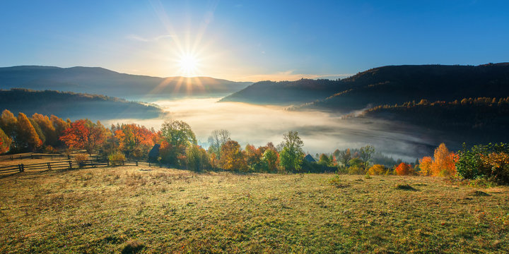 amazing countryside in fall season at sunrise. gorgeous view in to the valley full of fog in morning light. sun above the distant mountain. fence through rural field on the hillside. beautiful autumn