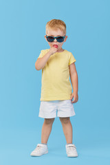 Little serious fashionable toddler boy in summer clothes and trendy sunglasses. Eats sweet candy on stick Isolated on blue background