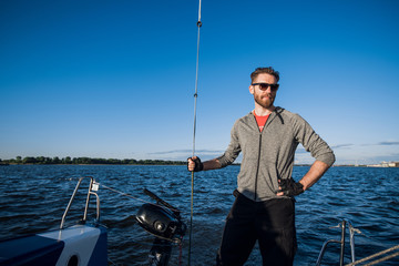 Obraz premium Yacht captain with a beard stands on sail boom on a sailing yacht, holding the rope in his hand and smiling, feeling happy. Adult yachtsman travelling around the world. Copy space