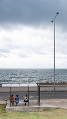 Obraz premium Three people waiting for the bus at a bus stop facing the sea, with a lighting pole