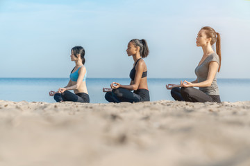 Side view shot of women making yoga meditation in lotus pose on sunny beach near water