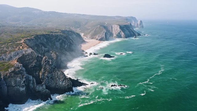 Aerial view of rocky cliff formation of Praia Grande beach, Portugal Atlantic coast. Cabo do Paco Lighthouse in Background