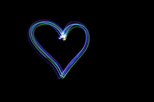Long exposure photograph of a heart shape outline in neon multi colour in an abstract parallel lines pattern against a black background. Light painting photography.