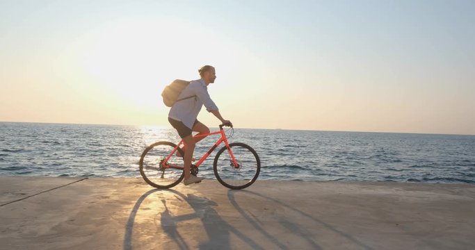 Young handsome male in casual wear ride on the colorful bicycle on the morning beach against beautiful sunset and the sea 