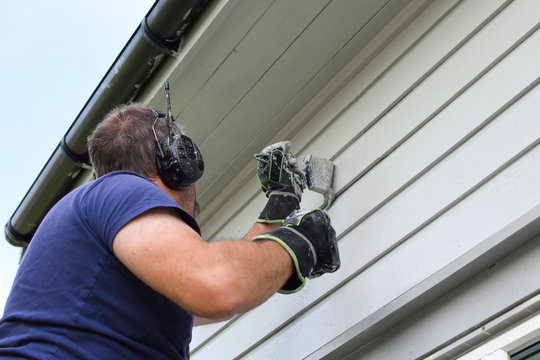 A man in headphones paints the exterior wall of a wooden house.