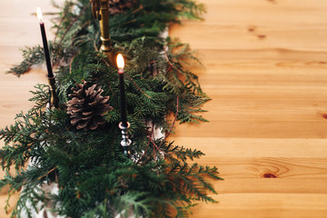 Stylish rustic christmas arrangement for festive dinner. Fir branches with pine cones and vintage candlestick with burning black candles on wooden table. Christmas rural table decor for feast