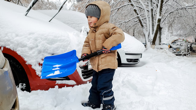 Image of boy in jacket helping to clean up the snow covered car after blizzard using big blue shovel