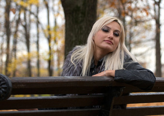 2017.10.21, Moscow, Russia. A young attractive woman wearing fur black jacket holding book sitting on the bench in falling park. Walking around autumn park.