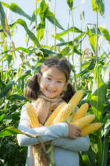Happy little girl holds ears of corn. Glad female child in casual clothes outdoor. Smiling kid in village. Girl with satisfied facial expression. Positive emotions, childhood idea