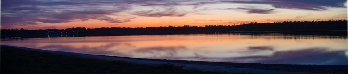 Fototapeta na wymiar Panoramic view on sunset at the lake romantic with smooth waves with shore in the foreground and forest behind