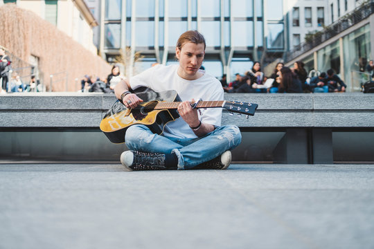white musician sitting on the floor in a urban square with stone floor