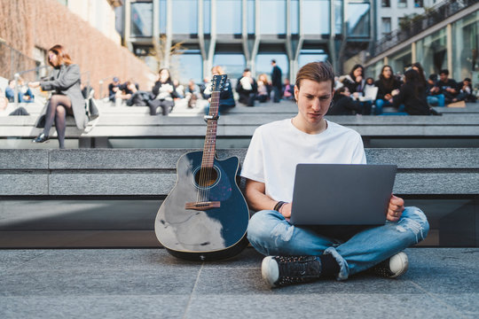 white man using laptop computer in square beside his classical guitar