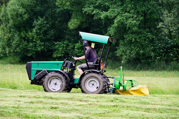 Man in shorts on small tractor mows grass in meadow with forest in background.